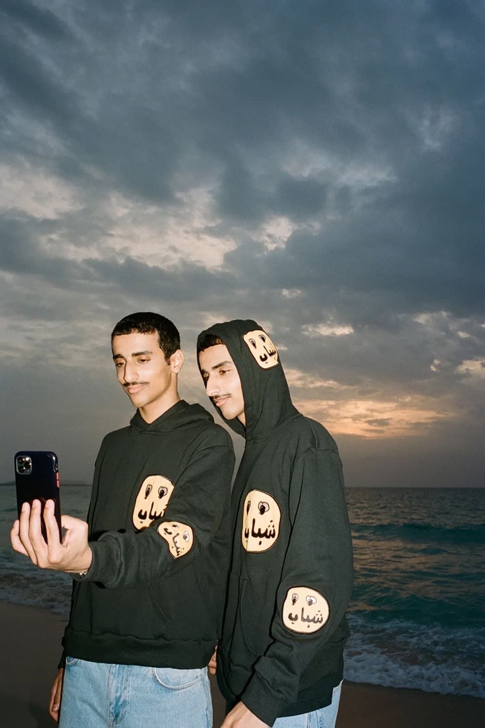 Two young men on a beach at dusk looking at a phone, in black hoodies with patches that read shabab in Arabic.