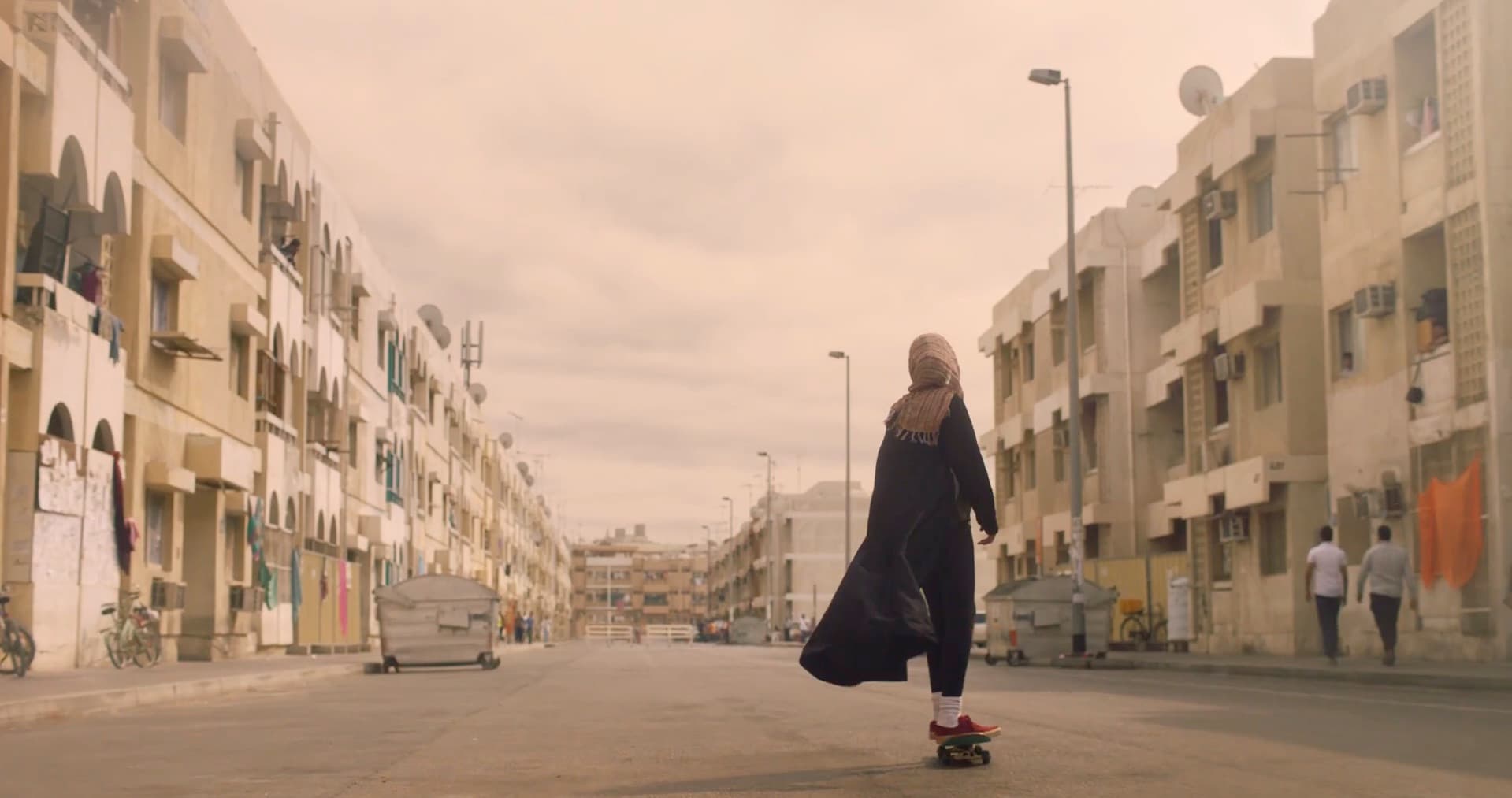 A young woman in an abaya and headscarf riding a skateboard down an empty street between apartment blocks.